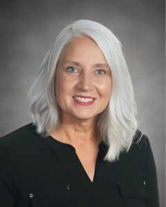 A portrait photo of Missy Testerman. She has white hair, fair skin, and blue eyes. She's wearing a black v-neck blouse and pink lipstick.